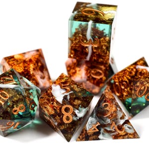 Op de afbeelding: Een verzameling polyhedrale dobbelstenen met een uniek ontwerp. De dobbelstenen zijn transparant met goudfolie en koperkleurige cijfers. Sommige dobbelstenen hebben een turquoise of wit wervel-effect. De dobbelstenen zijn gerangschikt op een witte ondergrond.