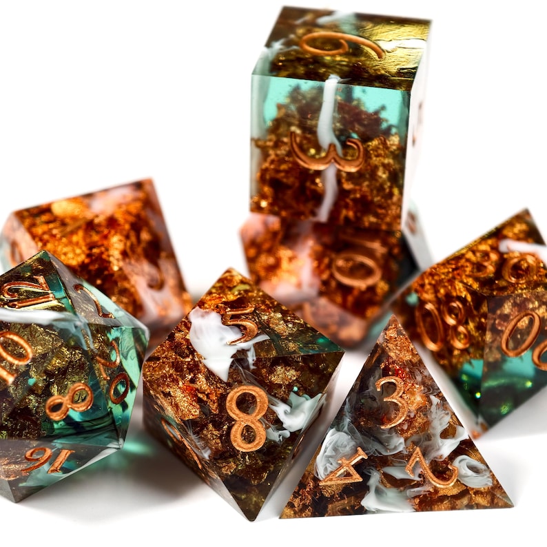 Op de afbeelding: Een verzameling polyhedrale dobbelstenen met een uniek ontwerp. De dobbelstenen zijn transparant met goudfolie en koperkleurige cijfers. Sommige dobbelstenen hebben een turquoise of wit wervel-effect. De dobbelstenen zijn gerangschikt op een witte ondergrond.