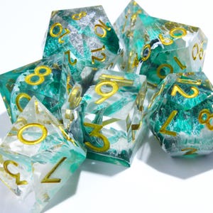 Op de afbeelding: Een verzameling polyhedrale dobbelstenen met een heldere harsbasis, gemengd met tinten groen en wit. De cijfers zijn goudkleurig. De dobbelstenen zijn ontworpen voor tabletopspellen.
