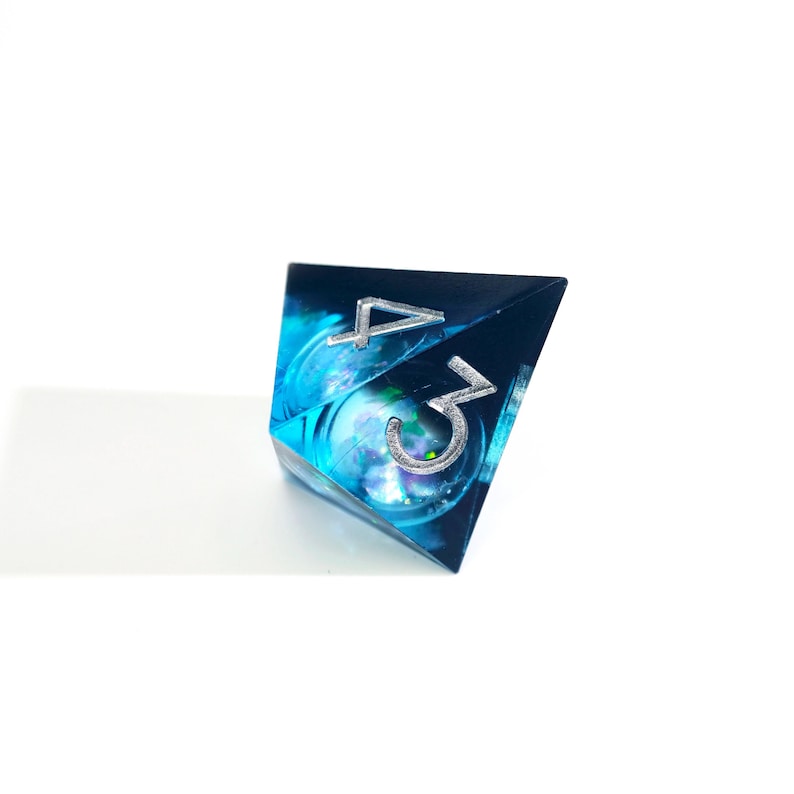 May include: A blue and teal polyhedral die with the numbers 3 and 4 in silver. The die has a swirling, galaxy-like design inside. The die is set against a white background.