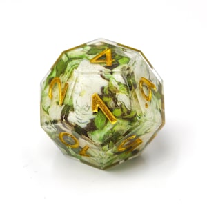 K&ouml;nnte beinhalten: Ein mehrseitiger polyedrischer W&uuml;rfel aus klarem Harz mit einem floralen Muster aus wei&szlig;en und gr&uuml;nen Bl&auml;ttern im Inneren. Die Zahlen sind in die Oberfl&auml;che eingraviert und mit einem metallisch goldenen Pigment gef&uuml;llt, das sich vom Design abhebt.