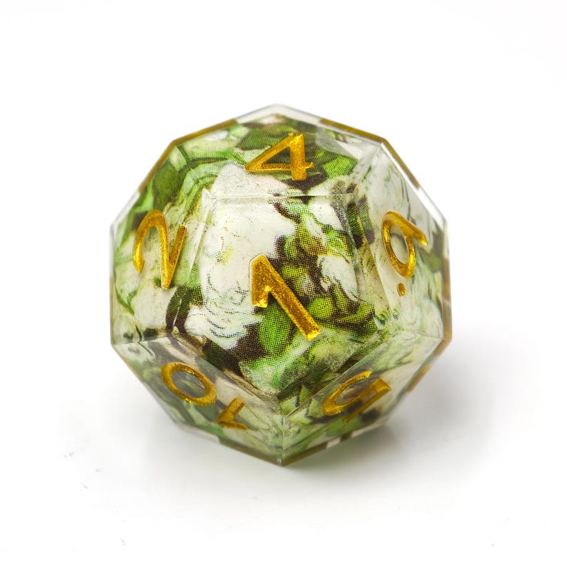 K&ouml;nnte beinhalten: Ein mehrseitiger polyedrischer W&uuml;rfel aus klarem Harz mit einem floralen Muster aus wei&szlig;en und gr&uuml;nen Bl&auml;ttern im Inneren. Die Zahlen sind in die Oberfl&auml;che eingraviert und mit einem metallisch goldenen Pigment gef&uuml;llt, das sich vom Design abhebt.