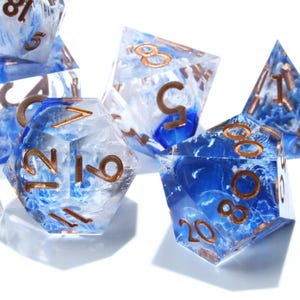 May include: A collection of polyhedral dice with a blue and white marbled effect. The dice have copper-colored numbers and are designed for tabletop gaming. The set includes various shapes, such as a d20, d12, and d6.
