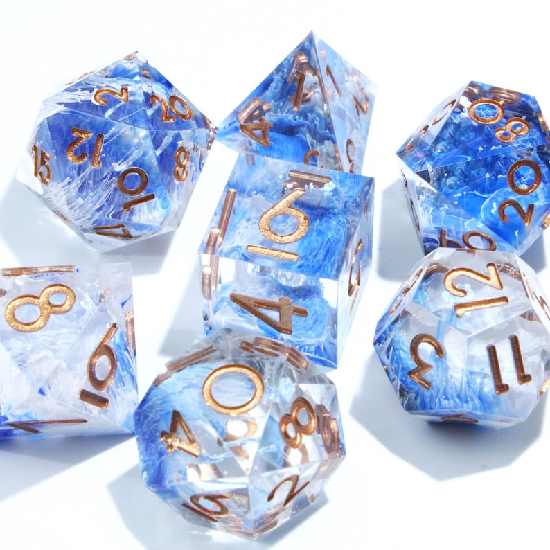 May include: A set of seven polyhedral dice with a clear resin base, blue wisps, and gold-colored numbers. The dice are in various shapes, including a d20, d12, d10, d8, d6, and d4, suitable for tabletop games.