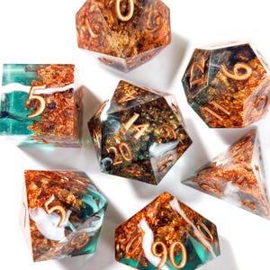 Op de afbeelding: Een verzameling polyhedrale dobbelstenen in verschillende vormen. De dobbelstenen hebben een mix van teal, goud en witte kleuren. De cijfers zijn koperkleurig. De dobbelstenen zijn voor tabletop games.