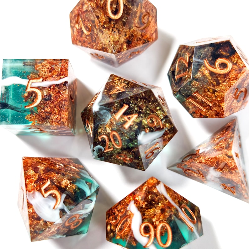 Op de afbeelding: Een verzameling polyhedrale dobbelstenen in verschillende vormen. De dobbelstenen hebben een mix van teal, goud en witte kleuren. De cijfers zijn koperkleurig. De dobbelstenen zijn voor tabletop games.