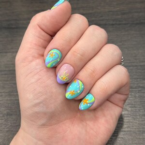 May include: A hand displaying a set of painted nails. The nails are oval-shaped and feature a design with a light blue base, swirls of green and purple, and orange flower accents. Some nails have white polka dots.