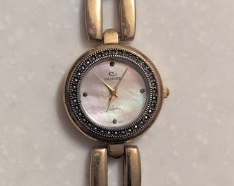 May include: A gold-toned quartz watch with a mother-of-pearl face and a decorative bezel. The watch features a gold-toned band with rectangular links and decorative accents. The watch face has the brand name "Cenere" and the word "Quartz" printed on it.