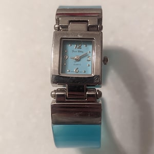 May include: A silver-toned Fred Belay quartz watch with a square, light blue face. The watch has a matching light blue band. The watch face displays the numbers 12, 3, 6, and 9, with small dots marking the other hours.