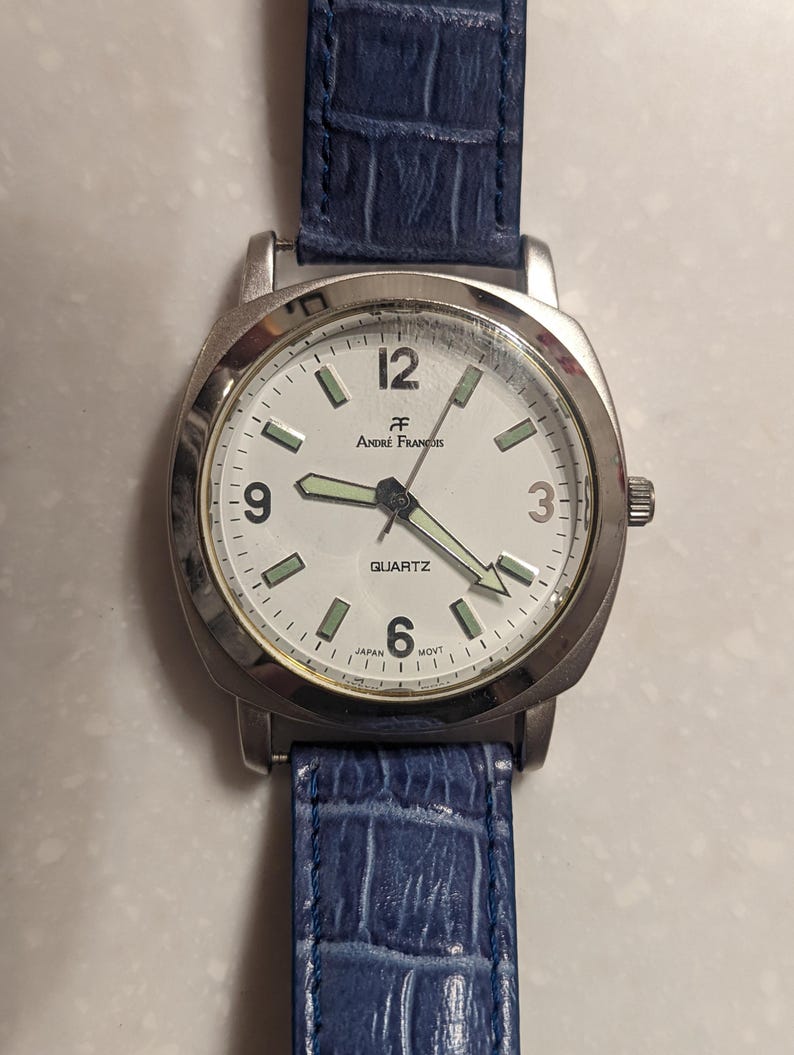 May include: A silver Andr&eacute; Fran&ccedil;ois quartz watch with a white face, black numerals, and luminous hands. The watch has a blue leather strap with a textured pattern. The watch face reads "QUARTZ" and "JAPAN MOVT".