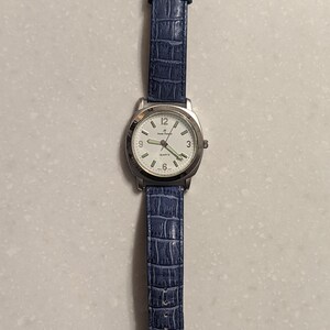 May include: A silver wristwatch with a white face and a blue leather strap. The watch has a gold-coloured buckle and displays the time with black numerals. The strap has a textured, crocodile-like pattern.