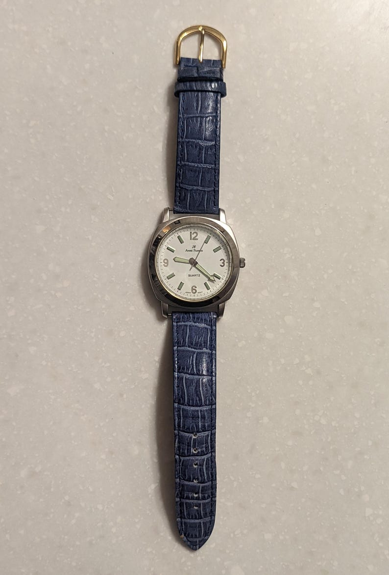 May include: A silver wristwatch with a white face and a blue leather strap. The watch has a gold-coloured buckle and displays the time with black numerals. The strap has a textured, crocodile-like pattern.