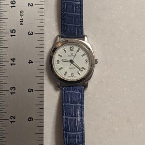 May include: A silver Anne Klein quartz watch with a white face, black numerals, and a blue faux-leather strap. The watch has a gold-coloured buckle and is approximately 20 cm long.