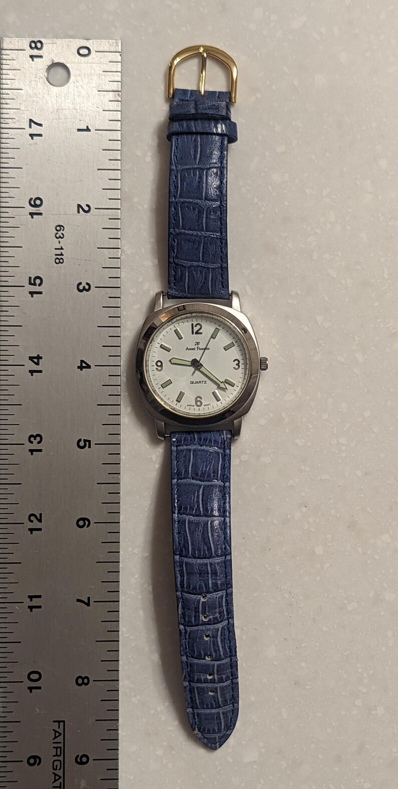May include: A silver Anne Klein quartz watch with a white face, black numerals, and a blue faux-leather strap. The watch has a gold-coloured buckle and is approximately 20 cm long.