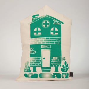 May include: A cream-colored pillow with a green illustration of a building. The building has a door, windows, and the word "EDIFICIO" above the door. The pillow has a tag on the bottom right corner.
