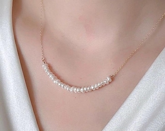 Puede incluir: Un delicado collar de oro con una fila de pequeñas perlas blancas. Las perlas están ensartadas juntas, creando una curva sutil y elegante. El collar se muestra sobre un fondo blanco, resaltando la simplicidad y sofisticación de la joya.