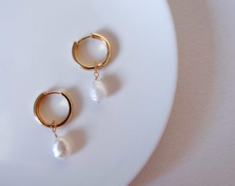 Puede incluir: Un par de pendientes de aro dorados con una perla blanca colgante. Las perlas tienen una forma irregular y orgánica. Los pendientes están sobre una superficie blanca, añadiendo un toque de elegancia natural.
