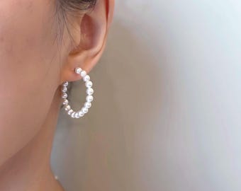 May include: A close-up of a pearl hoop earring. The earring is a half-circle shape, composed of small, round, white pearls. The earring is worn on the ear of a person, with the ear and part of the face visible.
