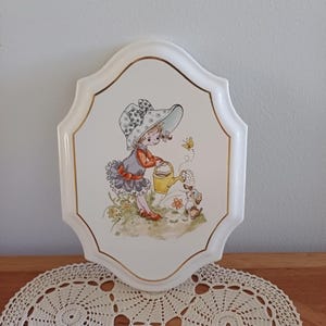 May include: A decorative wall plaque with a picture of a girl watering a puppy with a yellow watering can. The plaque has a white border with gold trim and a scalloped edge. The girl wears a hat and dress. A butterfly is also present.