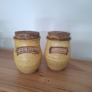 May include: Two speckled yellow ceramic canisters with cork lids. Each canister features a raised rectangular plaque with embossed text. One plaque reads TEA BAGS and the other reads COFFEE. The containers have a rounded shape and a textured finish.