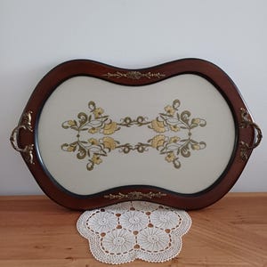 May include: An oval serving tray with a dark brown wooden frame and brass handles. The tray's interior features a cream-coloured surface with a floral design in yellow and green. A decorative doily sits beneath the tray.