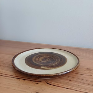 May include: A ceramic plate featuring a cream-coloured outer ring and a brown, spiral-patterned centre. The plate has a dark brown rim and is placed on a wooden surface. This is a decorative item.