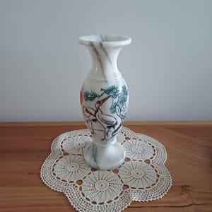 May include: White marble vase with a flared top and base, featuring a painted design of cranes and foliage in black, red, and blue. The vase is set on a cream-colored, crocheted doily, with a wooden surface in the background.