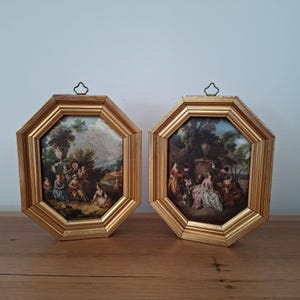 May include: Two octagonal wall art pieces featuring gold-toned frames and metal hanging loops. Each frame contains a detailed landscape painting depicting groups of people in period clothing gathered outdoors near stone urns and lush greenery.