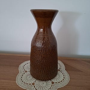 May include: A tall, cylindrical brown ceramic vase with a flared rim and a narrow neck. The lower body of the vase features a vertical, textured, bark-like pattern, while the upper neck and rim have a smooth, glossy, dark brown glazed finish.
