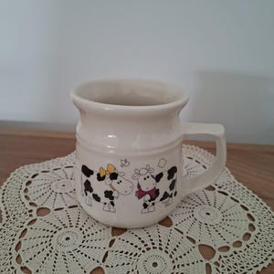 May include: A cream-coloured ceramic mug featuring a graphic illustration of two black and white cows. One cow wears a yellow bow, and the other wears a dark pink bow. The mug has a wide, rounded body with a sturdy handle and a smooth, glossy finish.