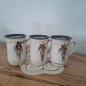 May include: Three off-white ceramic mugs with a dark blue rim and a horizontal brown line encircling the body. Each mug features a hand-painted botanical design of brown and blue leaves. The mugs have a sturdy handle and a cylindrical shape.