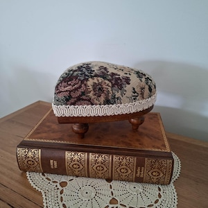 May include: A heart-shaped pincushion with a floral tapestry pattern in shades of brown, cream, and green, featuring a cream-coloured decorative trim and small wooden feet, resting on a brown leather-bound book titled Oliver Twist by Charles Dickens.