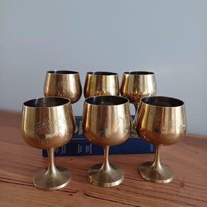 May include: A set of six vintage brass wine goblets. Each goblet has an etched floral design and a rounded base. The goblets are displayed on a wooden surface, with a book visible in the background.