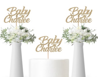 May include: Three gold glitter cake toppers with the text "Baby Charlie" on each. The toppers are on a white cake, a white vase with white flowers, and another white vase with white flowers.