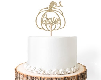 May include: Gold glitter pumpkin cake topper with the name Grayson. The topper is on a white cake with white frosting and a wood cake stand.