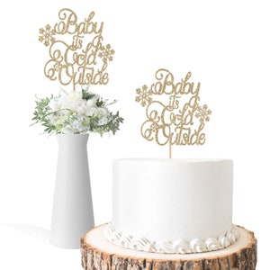 May include: Two gold glitter cake toppers with the text "Baby It's Cold Outside". One topper is on a white cake with white frosting and the other is on a white vase with a bouquet of white flowers.