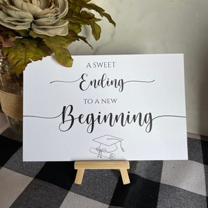 May include: A white card with black text that reads "A Sweet Ending to a New Beginning". A graduation cap and diploma are drawn below the text. The card is on a small wooden easel.
