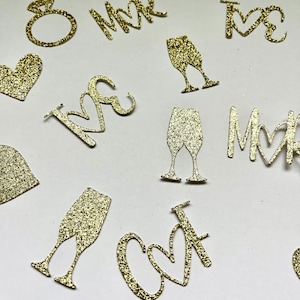 May include: Gold glitter cutouts of wedding-themed shapes, including a ring, champagne glasses, hearts, and the words "Mr" and "Mrs". The cutouts are scattered on a white surface, ideal for wedding decorations.