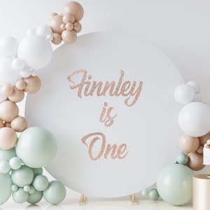 May include: A white round backdrop with the text "Finley is One" in rose gold glitter. The backdrop is decorated with a garland of white, beige, and light green balloons.