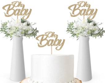 May include: Three gold glitter 'Oh Baby' cake toppers. Two toppers are in white vases with white flowers. The third topper is on a white cake with white frosting.
