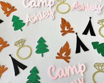 May include: Assorted glittery die-cut shapes including campfires, pine trees, diamond rings, and black tents. The words "Camp Ashley" are also present in pink glitter. These decorative elements are ideal for crafting or party decorations.