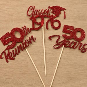 May include: Two red glitter cake toppers on wooden sticks. One topper reads Class of 1976 with a graduation cap icon. The other topper reads 50th Reunion and 50 Years in a cursive and block font combination. The glitter texture is visible throughout.