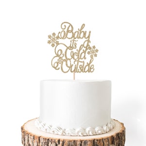 May include: A white cake with gold glitter frosting and a gold glitter cake topper that says "Baby it's cold outside".