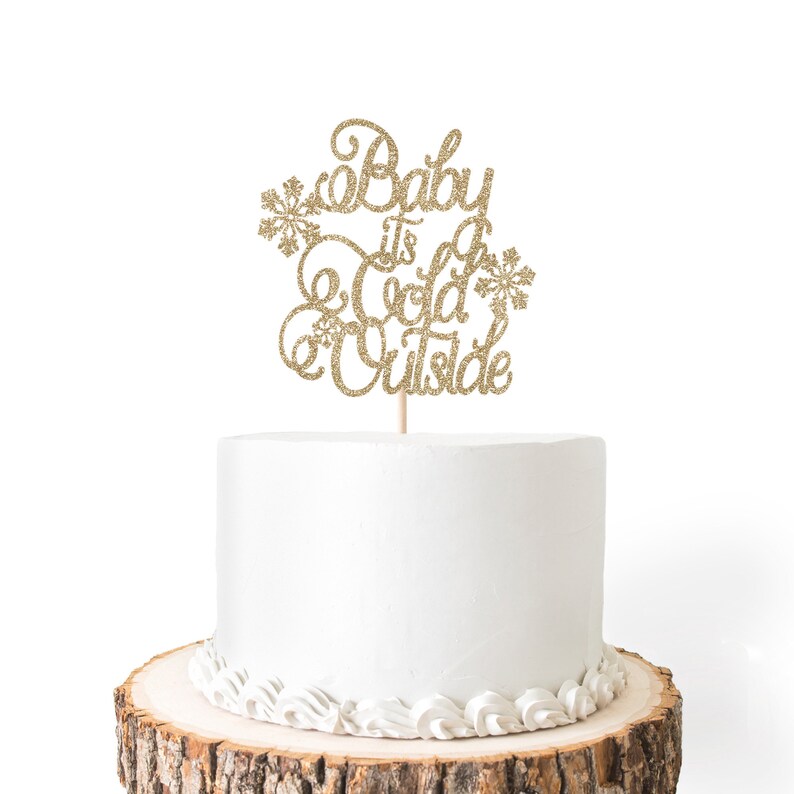 May include: A white cake with gold glitter frosting and a gold glitter cake topper that says "Baby it's cold outside".
