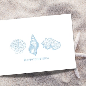 Op de afbeelding: Een witte wenskaart met drie blauwe schelpenillustraties en de tekst "HAPPY BIRTHDAY". Een witte zeester ligt op het zand naast de kaart. De kaart heeft een strandthema.