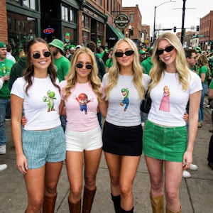May include: Four young women wearing sunglasses and graphic t-shirts pose outdoors. The shirts feature characters from the Super Mario Bros. series: Yoshi, Mario, Luigi, and Princess Peach. They are standing in front of a brick building with a crowd in the background.