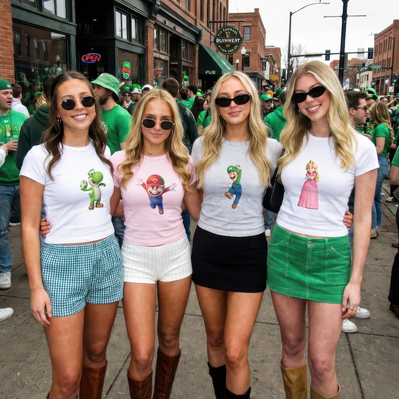 May include: Four young women wearing sunglasses and graphic t-shirts pose outdoors. The shirts feature characters from the Super Mario Bros. series: Yoshi, Mario, Luigi, and Princess Peach. They are standing in front of a brick building with a crowd in the background.