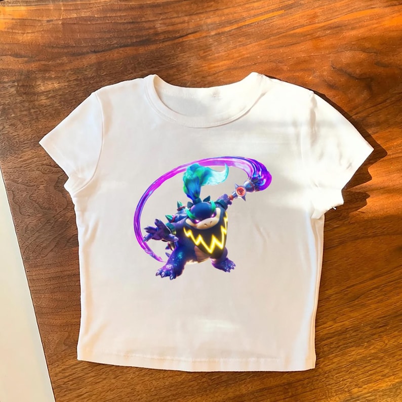 May include: White crop top featuring a colorful graphic of a cartoon character with a blue and green mohawk, holding a weapon with a purple energy trail. The character has a yellow lightning bolt design on its chest.