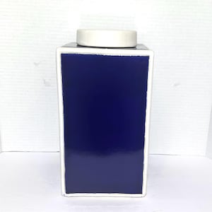 May include: A rectangular ceramic canister featuring a deep, glossy navy blue finish on its front and side panels, framed by a crisp white border. The container is topped with a matching white, flat-topped lid that sits flush with the top edge.