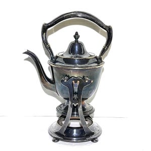 May include: A silver-plated tea kettle with a curved spout, a domed lid, and a high arched handle. The kettle is mounted on a matching metal stand with three feet, featuring a central circular opening designed to hold a heat source underneath.